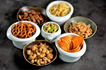 Different kinds of snacks - chips, salted peanuts, cashews, almonds and pistachios, pretzels with salt, potatoes, salted straw.