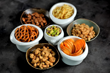 Different kinds of snacks - chips, salted peanuts, cashews, almonds and pistachios, pretzels with salt, potatoes, salted straw.