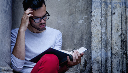 Fototapeta premium Portrait handsome bearded man wearing glasses. Young male student reading book and relaxing in old European city.