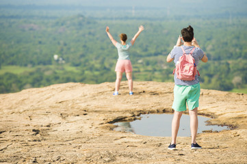 A boy is taking a photo of his girlfriend on top of a mountain near Sigiriya, Sri Lanka. Blurred green vegetation in the background.