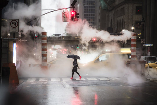 A woman with an umbrella and red high heels shoes is crossing the 42nd street in Manhattan. Taxi and steam coming out from from the manholes in the background. New York City, Usa.