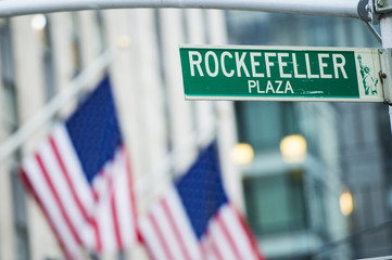 Close-up view of green street sign depicting it is Rockefeller Plaza in Midtown Manhattan, New-York. Blurred American flags in the background © Travel Wild