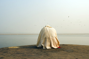 A holy man dressed in white is praying and meditating on one of the many Ghats of Varanasi in front...