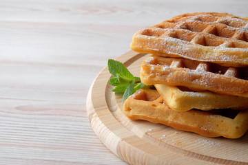 A pile of Belgian waffles sprinkled with powdered sugar on a light cutting board. Closeup view.