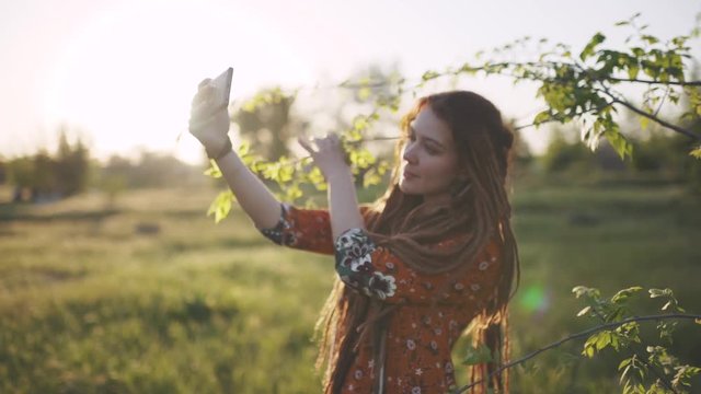 attractive hippie woman with dreadlocks making selfie on smartphone at sunset near glade