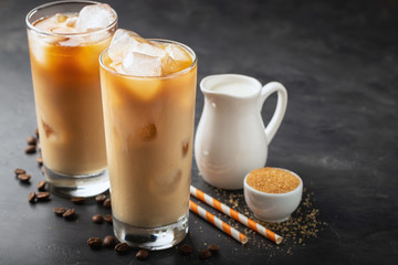 Ice coffee in a tall glass with cream poured over, brown sugar and coffee beans. Cold summer drink on a dark background. With copy space