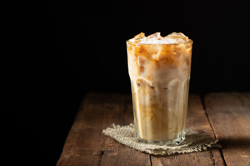Ice coffee in a tall glass with cream poured over and coffee beans on a old rustic wooden table. Cold summer drink on a dark wooden background with copy space