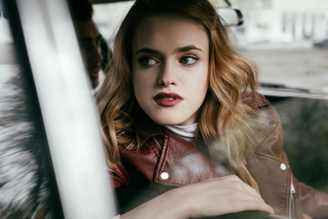 Fototapeta na wymiar beautiful young woman in leather jacket looking away while sitting in car