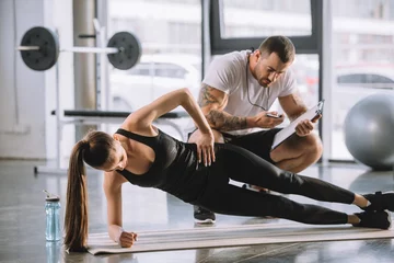 Tuinposter Male personal trainer looking at timer and young athletic woman doing side plank on fitness mat © LIGHTFIELD STUDIOS