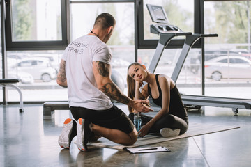 Fototapeta na wymiar male personal trainer explaining talking to young athletic woman sitting on fitness mat at gym