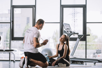 Fototapeta na wymiar male personal trainer showing timer to young athletic woman sitting on fitness mat with bottle of water at gym