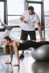 Obraz na płótnie Canvas male personal trainer with timer and young athletic woman doing push ups on fitness ball at gym
