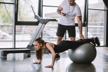 male personal trainer with timer and young athletic woman doing push ups on fitness ball at gym