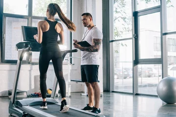 Tuinposter male personal trainer using timer while sportswoman running on treadmill at gym © LIGHTFIELD STUDIOS