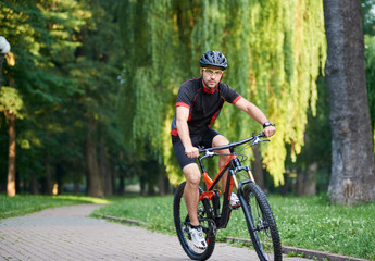 Sporty guy bicyclist in cycling clothes and protective helmet looking to camera while riding bike down park alley on summer day. Exercising, reaching goal