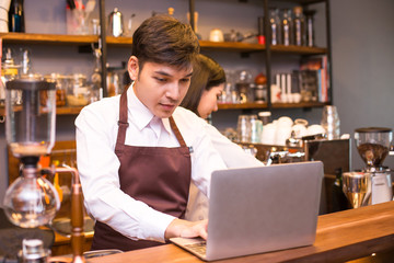 Asian male barista using laptop for work at coffee shop counter. Man with small business or sme concept.