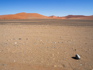 Fototapeta na wymiar Lonely egg shape rock stone and shadow on vast empty dried desert landscape with natural sand dune curved ridge and clear blue sky background, Namib desert