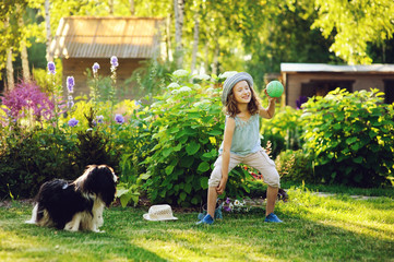 happy child girl playing with her spaniel dog and throwing ball, enjoying sunny summer day in garden