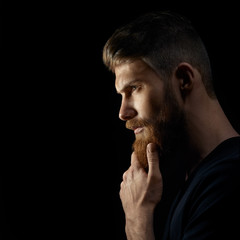 Attractive pensive young man looks into the distance stroking his beard over black background