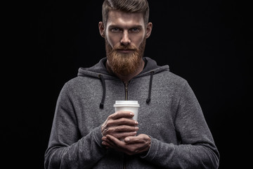 Dramatic shot of handsome young bearded man in gray hoodie having coffee Male taking rest and drinking fresh morning coffee Young hipster bearded man with coffee in hand Template mockup copy space.