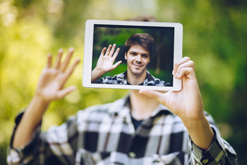 detail shot of a handsome young man taking a selfie with a tablet, smiling and waving at the camera.