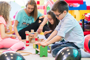 Cute pre-school boy cooperating with his colleagues at the construction of a structure made of wooden toy blocks, under the guidance of a young kindergarten teacher