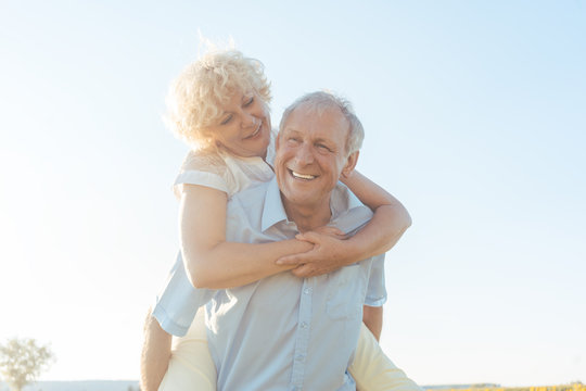 Low-angle View Portrait Of A Happy Senior Man Laughing, While Carrying His Partner On His Back In A Sunny Day Of Summer In The Countryside