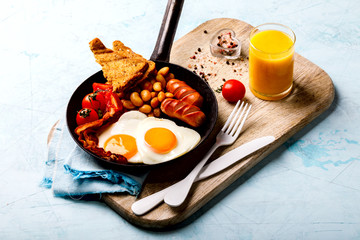 Traditional English Breakfast in the Frying Pan Eggs Heart shape. Valentine's Day. Festive Food on the Blue Background.Sausages, Bacon, Beans,Toasts,Coffe and Orange juice
