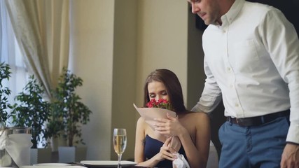 Beautiful woman is waiting for her boyfriend in restaurant, he is coming and giving her flowers, kissing her. Happy girl is smelling flowers and smiling with pleasure.