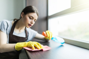 Young woman in yellow gloves holding rag and window cleaner in hands near the window indoors
