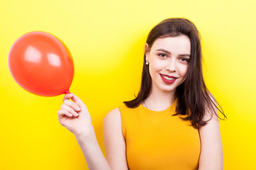 Smiling young woman playing with a red balloon on yellow background in studio