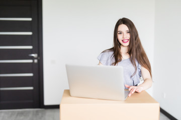 Beautiful woman moving in her new house and unpacking, she is sitting on the floor surrounded by boxes, using a laptop and smiling at camera