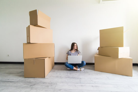 Young woman sitting on hardwood floor in new aprtmanet, using laptop next to cardboard box