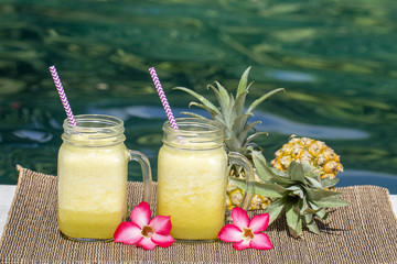 Homemade mango and pineapple smoothie made with coconut milk in two glass mug near swimming pool, close up. Refreshing tropical fruit drink