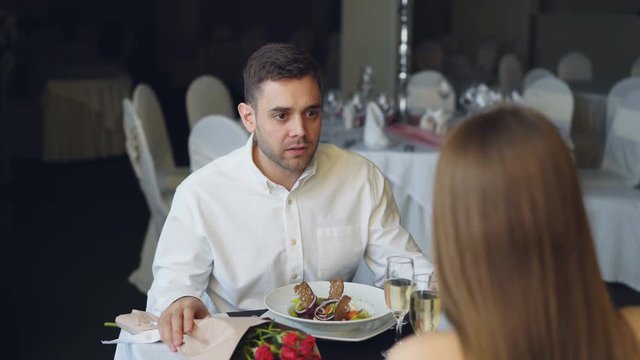 Young couple is quarreling while dining in restaurant, shouting and gesturing. Bouquet of roses, sparking champagne glasses and plates with food are visible.