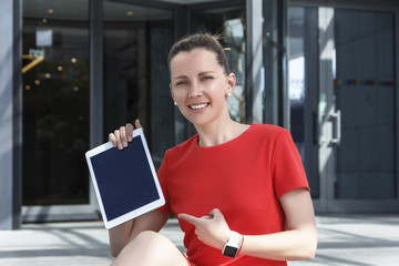 A young woman in a red business dress is smiling and looking directly at the camera, sitting on the steps opposite the entrance to the business center and showing something on the tablet.