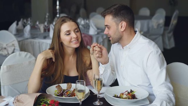 Happy loving couple is holding hands, talking and kissing during romantic dinner in restaurant. Affectionate relationship, love and fine dining concept.