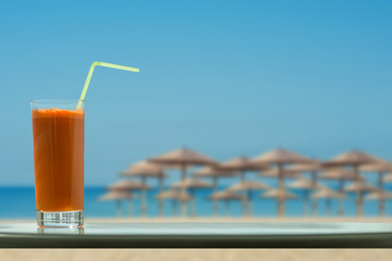A glass of carrot juice with a straw in the cafe on the beach and straw umbrellas backgrounds 