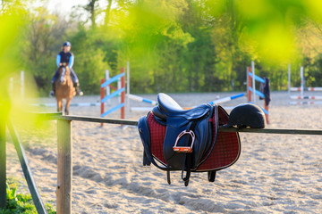 Horseback saddle on the wooden fence on the background of training rider in a sunny day