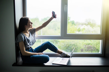 Happy woman in t-shirt sitting on the windowsill and making selfie on smartphone