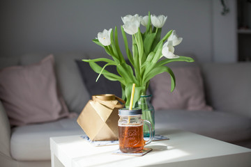 Interior. Room. A bouquet of tulips in a glass vase, a gold candlestick, a mug of lemonade on a white wooden table. Sofa, pillows. Spring. It's cozy. Living room.