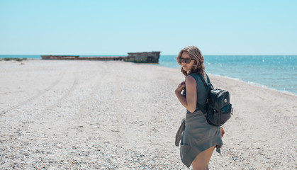 girl on a sea beach with a backpack and glasses in the background of a sunken ship