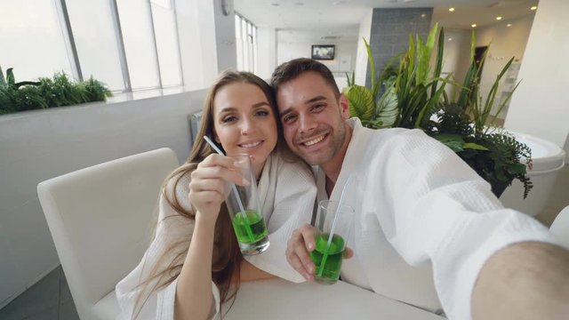 POV shot of happy lovers popular bloggers recording video for their followers while relaxing in spa salon. Young people in bathrobes are posing, laughing and kissing.