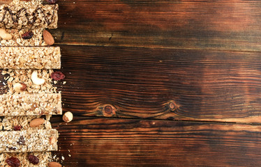 Variety of granola energy bars in row with scattered mixed nuts, cereals, cranberry on brown wood textured table background. Healthy nutritious vegan fitness food snack. Top view, copy space, close up