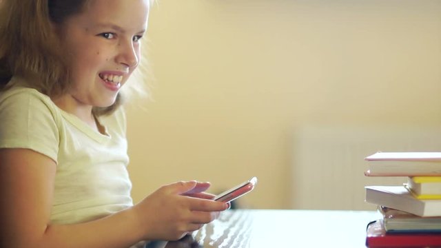 A sad girl is sitting over a pile of books. Angrily pushes them away and takes the phone. Internet addiction. He smiles cunningly. Using cell phone