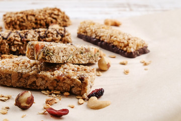 Various granola chocolate energy bars in row with scattered mixed nuts, cereals & dried fruit, grunged white wood table background. Healthy vegan fitness food snack. Top view, copy space, close up.