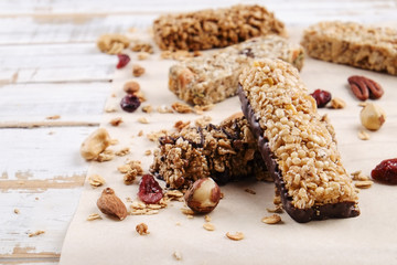 Various granola chocolate energy bars in row with scattered mixed nuts, cereals & dried fruit, grunged white wood table background. Healthy vegan fitness food snack. Top view, copy space, close up.