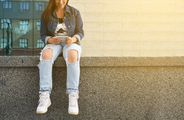 A beautiful girl in a jeans jacket, sitting on the steps on a city street, uses her smartphone. A woman communicates through the Internet. Lifestyle. Trend. Hipster.