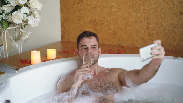 Young handsome man popular blogger is recording video in hot tub in day spa using smartphone. Burning candles, champagne glass and flowers are visible.