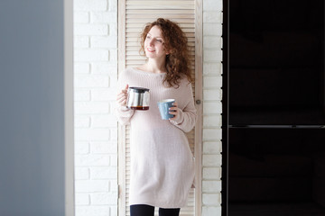 Beautiful caucasian woman with curly red hair drinking coffee,standing by the big window, enjoying a quiet cozy evening at home
